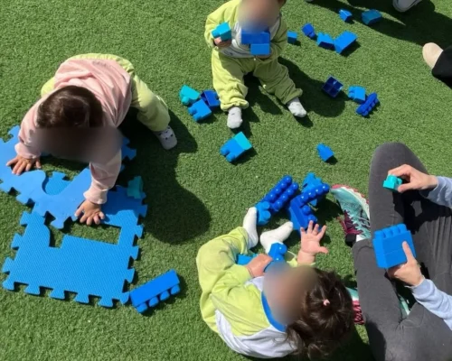 Niños jugando en un área al aire libre con bloques de construcción azules sobre césped artificial. Dos pequeños están sentados, uno manipulando piezas y otro sosteniendo bloques, mientras un adulto participa en la actividad.